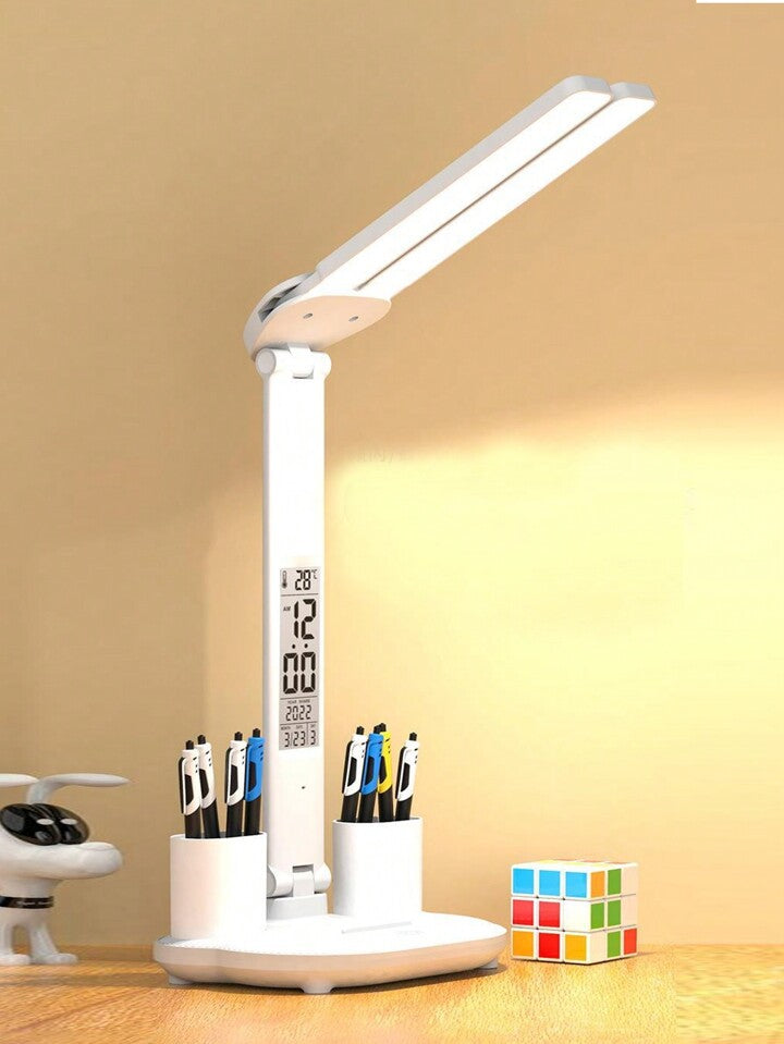 The LED Desk Lamp USB Rechargeable Dimmable Reading Light is a white desk lamp with eye protection and an adjustable arm. It features a built-in digital display showing time and temperature, includes pen holders on a round base stocked with assorted pens, has a colorful Rubik's cube nearby, and displays a small decorative airplane in the background.