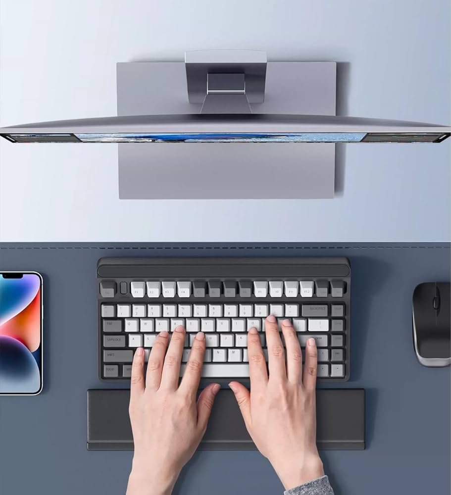 An overhead view shows someone typing on a compact mechanical keyboard with white and gray keys, accompanied by a curved monitor, smartphone, and wireless mouse on a light blue desk. The person's hands rest comfortably on the Keyboard Wrist Rest Pad, which provides ergonomic memory foam support for pain relief in an office setting.