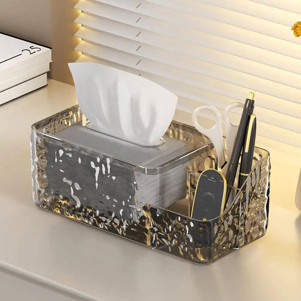 The Desktop Tissue Dispenser Stylish Tissue Holder is an elegant, transparent, textured container that organizes a tissue box, scissors, remote control, and pens on a desk next to window blinds. A notebook peeks into view on the left side.