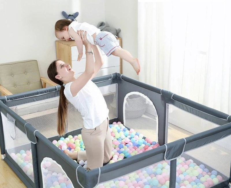 A woman joyfully lifts her baby above her head inside a Baby Activity Fence Playpen Safety Gate Children's Playground filled with plastic balls. The room, reminiscent of a children's playground, is bright, featuring a cozy sofa and sunlit window in the background.