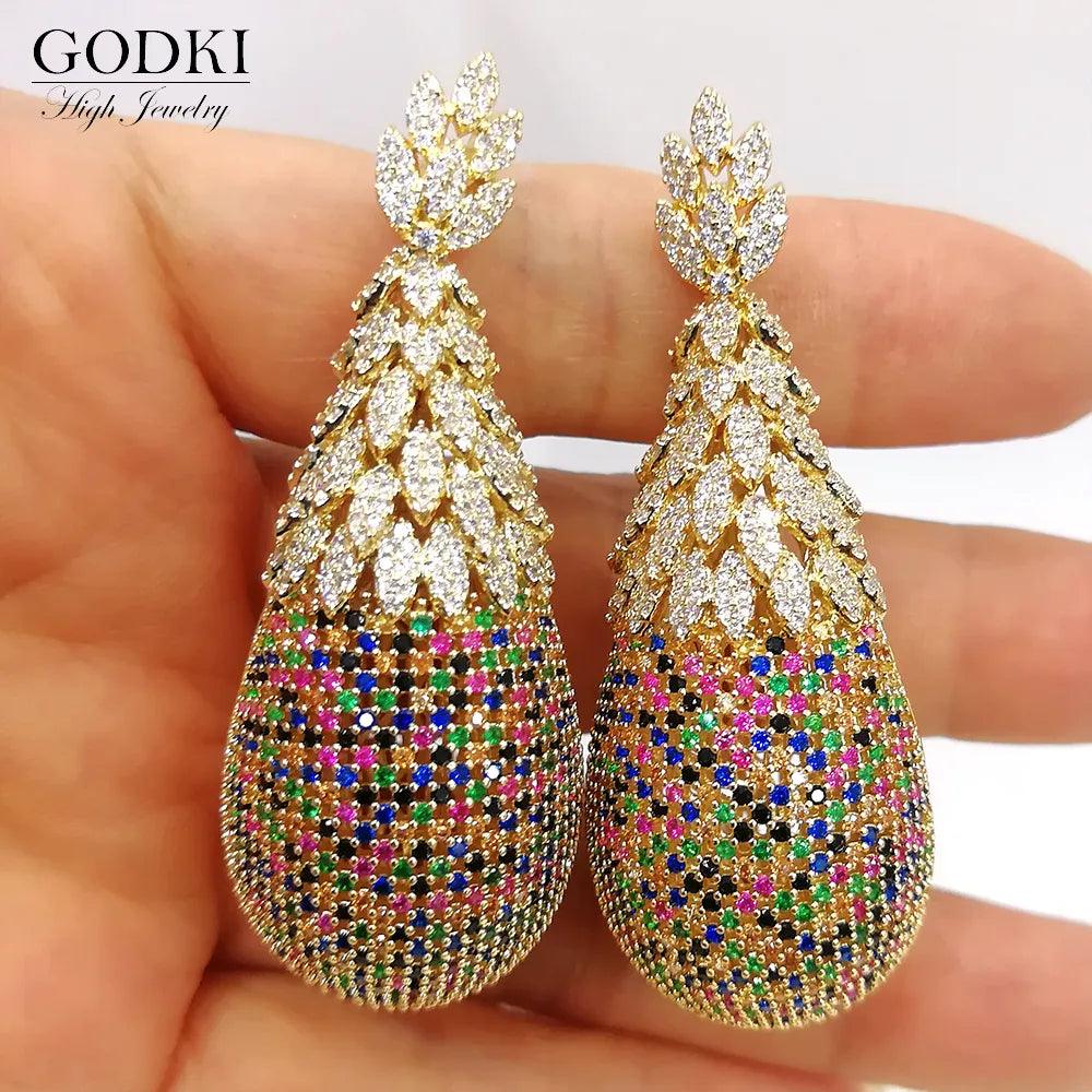 A pair of elaborate Pineapple Earrings Luxury Bridal Earrings, showcasing a gold-toned leaf motif at the top and a teardrop form embellished with multicolored gemstones, encapsulates the ornate charm of Dubai-style bridal jewelry.