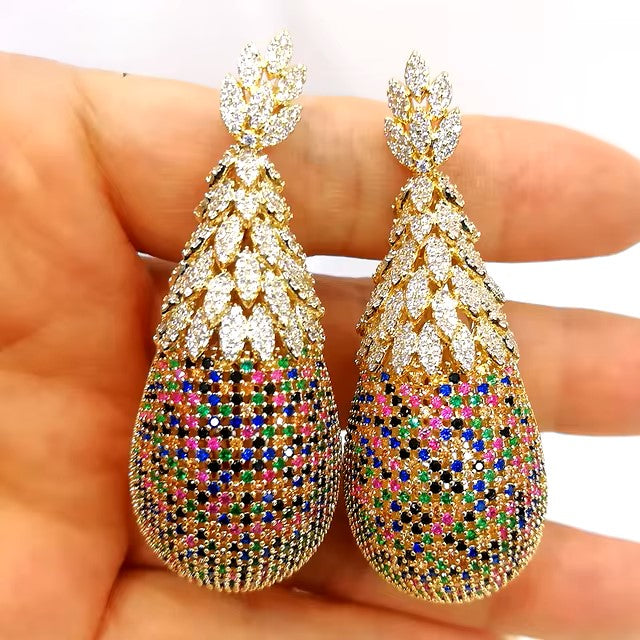 A pair of Pineapple Earrings Luxury Bridal Earrings, designed in a Dubai style with a sleek, leaf-like pattern on top, embellished in gold and adorned with multicolored and clear gemstones. They rest elegantly on a person's hand, highlighting their intricate detailing and sparkling surface, perfectly capturing the essence of luxurious bridal jewelry.