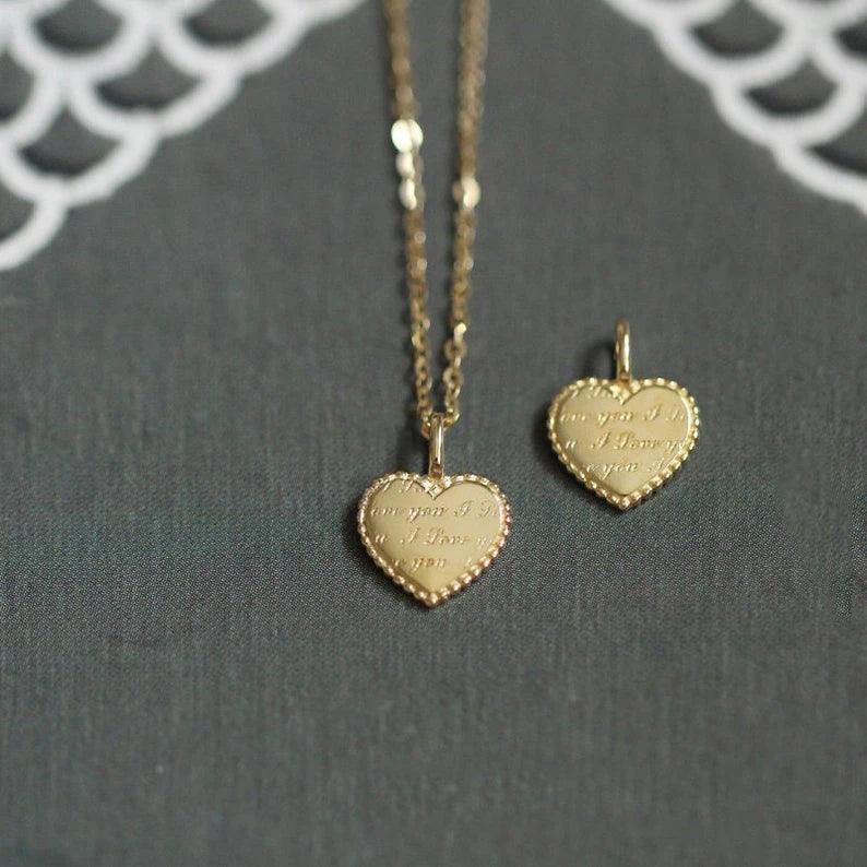 A close-up of a 9k solid gold heart charm engagement Victorian bride necklace and matching pendant on a dark fabric background with lace detailing. Both pieces, symbols of timeless romance, feature an engraved message on the surface.