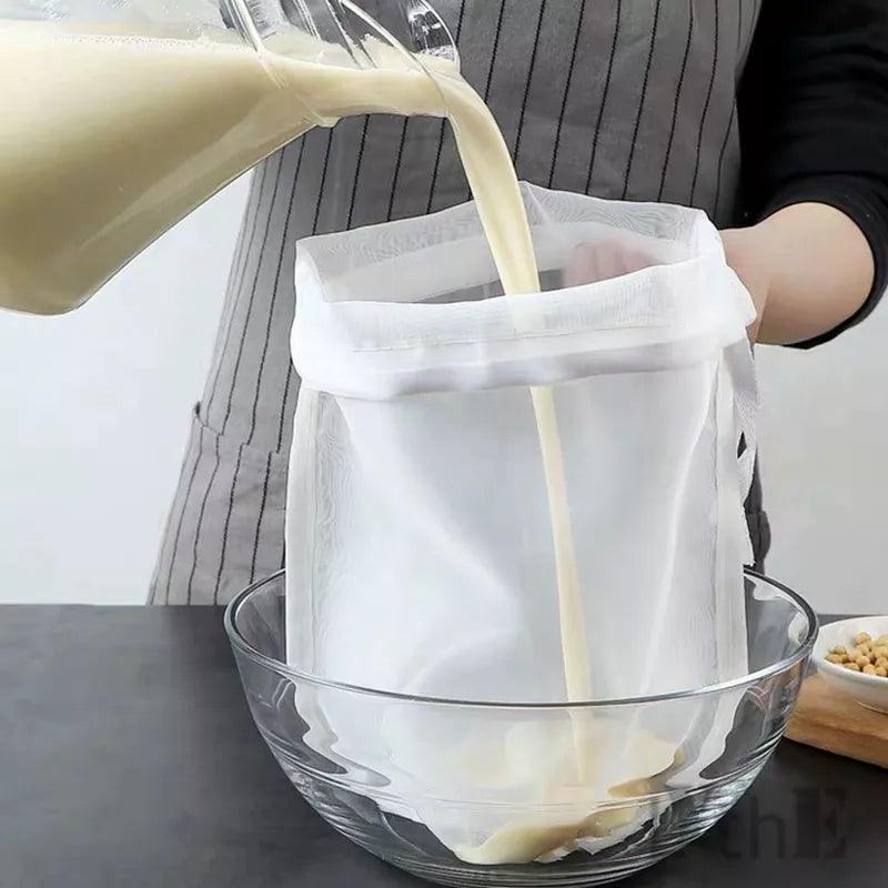 A person is expertly pouring a creamy liquid into the "Soy Milk Filter Bag," a versatile mesh kitchen tool, over a clear glass bowl. Clad in a gray apron with vertical stripes, they skillfully complete this nut milk filtering task, as nuts sit visibly in a nearby bowl.