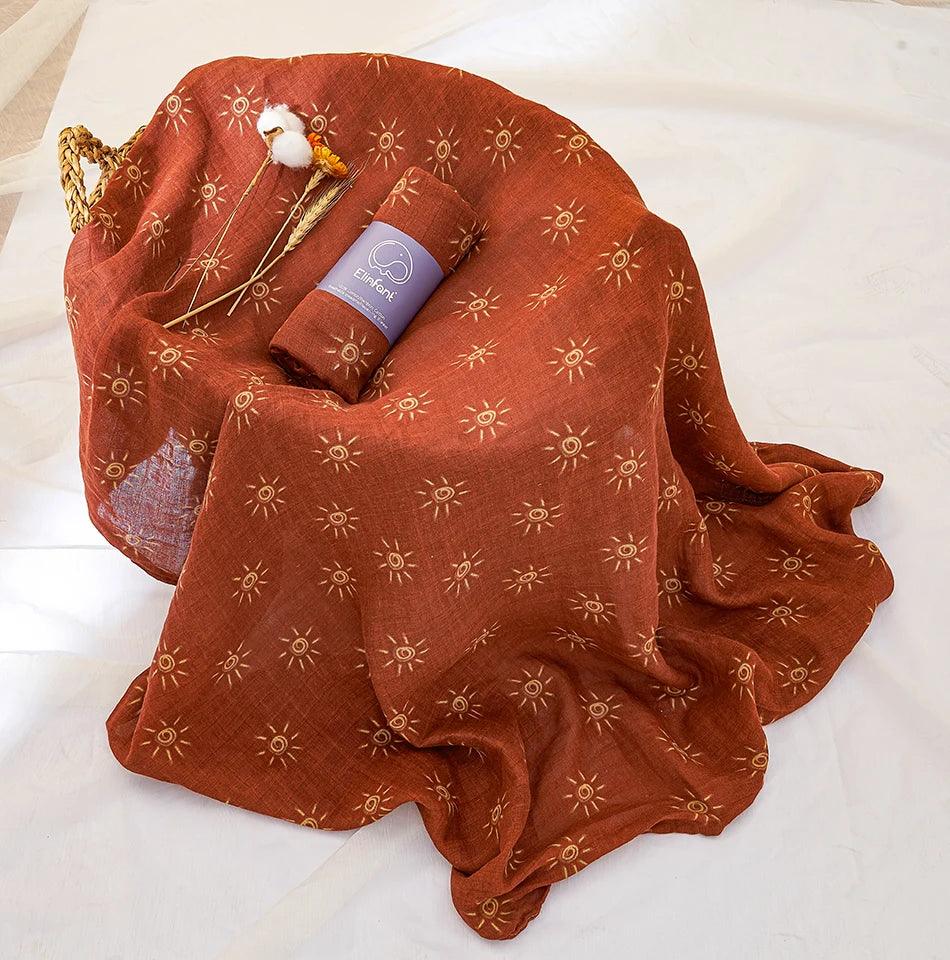 A rich brown organic cotton fabric featuring a sun pattern is gracefully draped over a basket. A rolled-up "Muslin swaddle blanket" and some dried flowers are artfully arranged on it, all set against a light-colored surface.