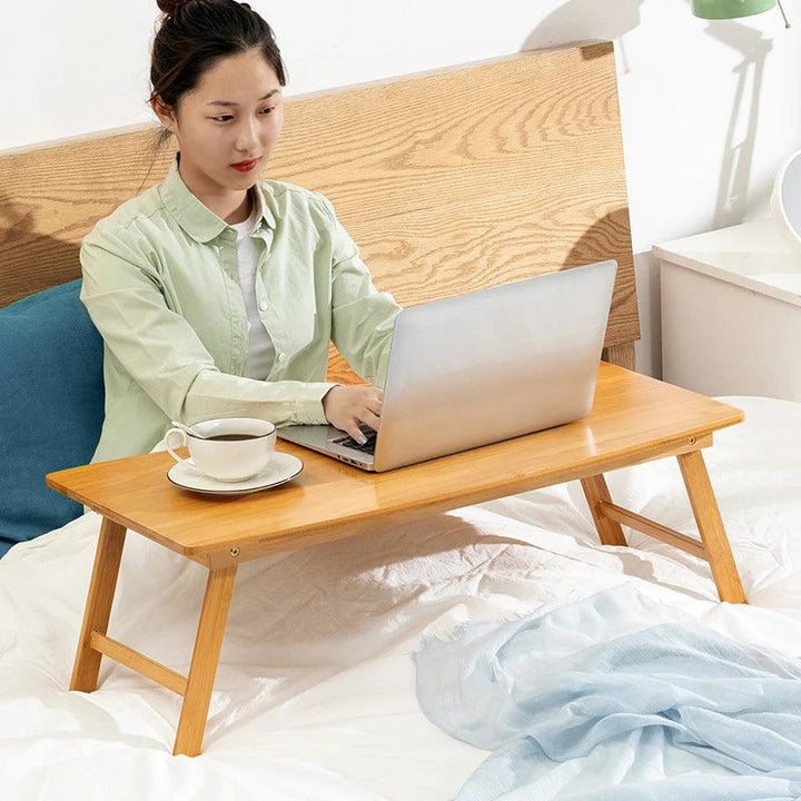 A woman sits on a bed with her laptop on the Portable Laptop Desk Bed Tray Table Adjustable Laptop Stand. A cup of coffee is placed next to her, and she remains focused on her work in a light green shirt. The room is adorned with white bedding and a wooden headboard, providing tranquility as she types away.