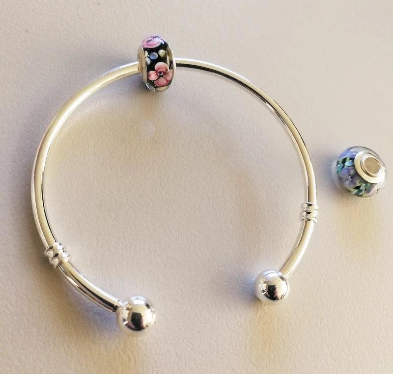 The Tibetan Silver Bangle European Charms Bracelet Jewelry Accessory comes with two detachable beads; one bead features pink and black floral patterns, while the other displays a blue and white floral design.