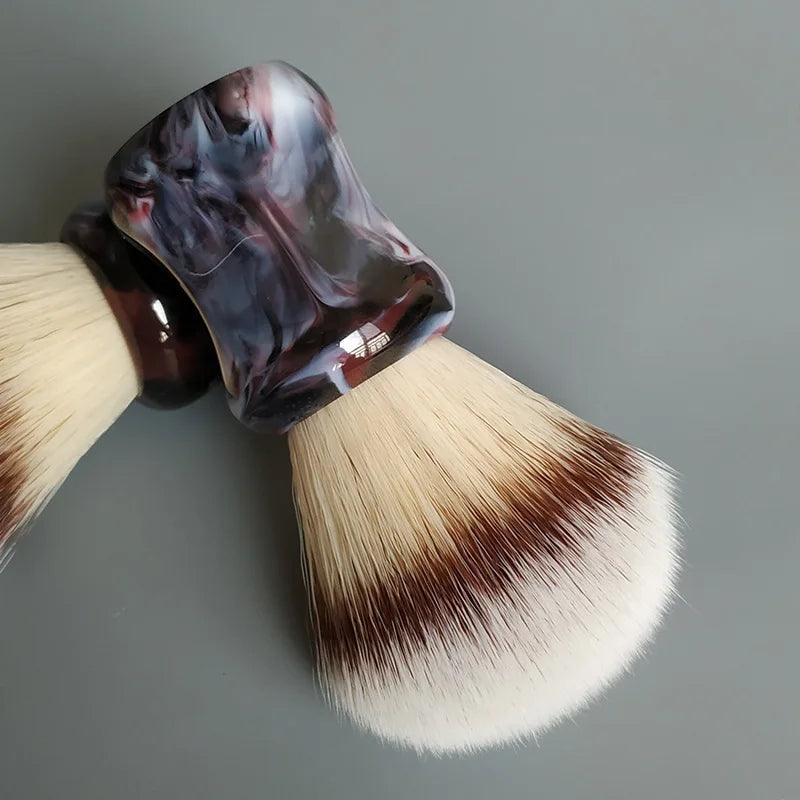 Close-up of two handmade shave brushes featuring light synthetic bristles and marbled dark and light resin handles, crafted for a refined shaving routine. These brushes, designed for professional-grade results, rest elegantly on a smooth surface.