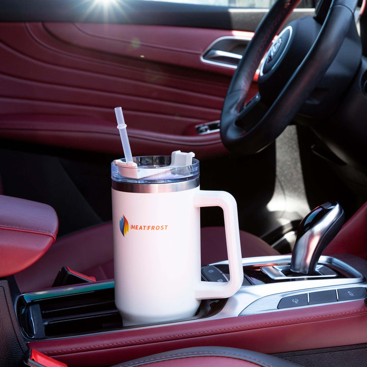 A 40oz Stainless Steel Tumbler Insulated Beverage Container, known as the Travel-Friendly Quencher, in white sits in the cup holder of a car with red leather seats and a shiny gear shift. Sunlight reflects off the quencher, creating a bright spot. This tumbler is an essential companion for drink enthusiasts.