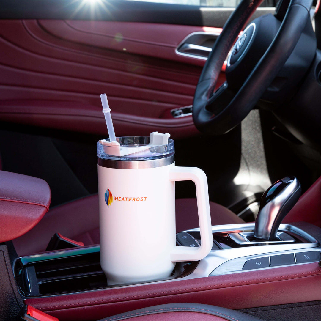 A 40oz white Stainless-Steel Tumbler Insulated Beverage Container sits in the cup holder of a red leather car interior, complete with a handle and straw. Sunlight reflects off the lid, highlighting the HEATFROST logo. This travel-friendly quencher is an essential companion for anyone who loves to enjoy their drinks at the perfect temperature.