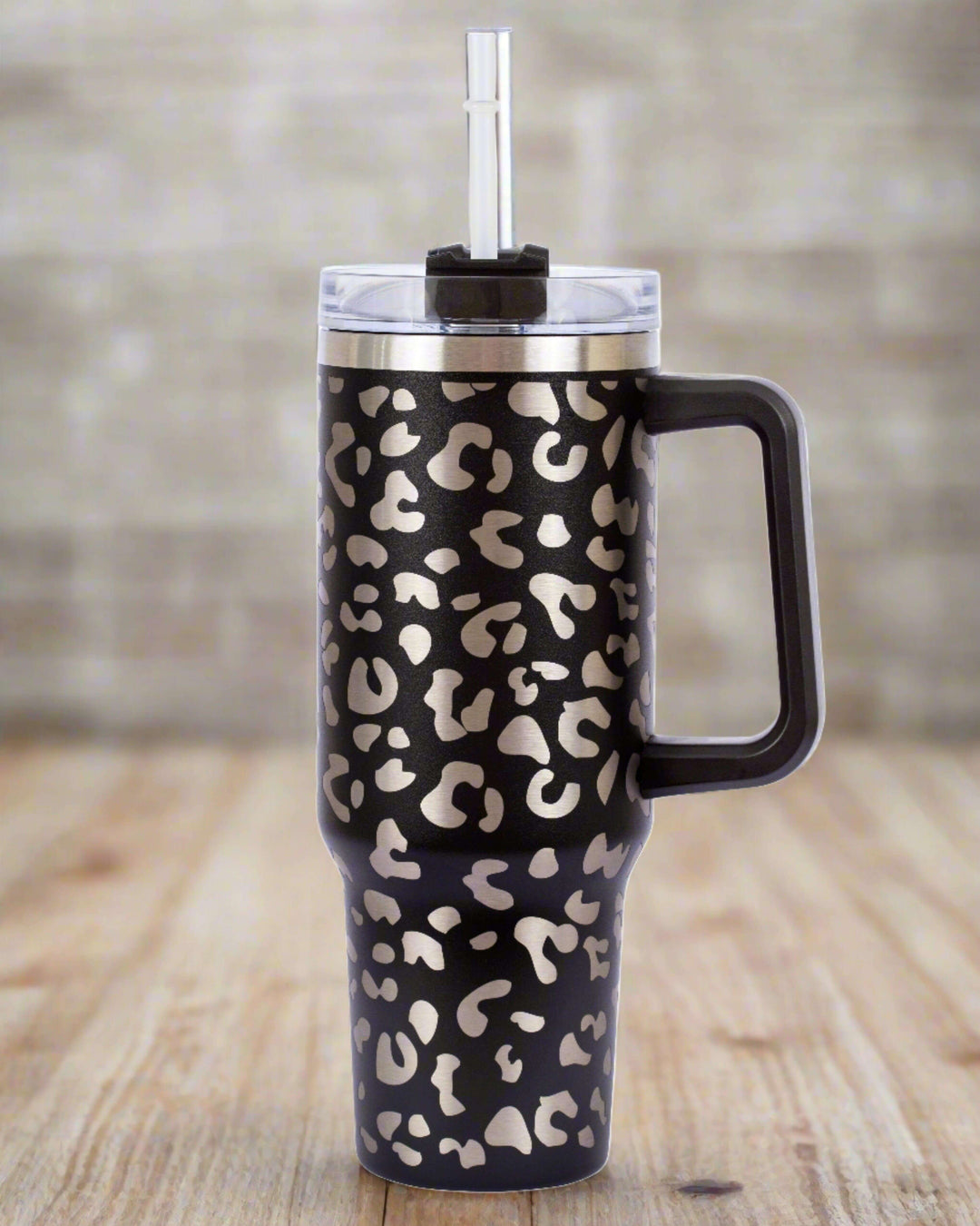 The 40oz Stainless Steel Tumbler Insulated Beverage Container, with its elegant silver leopard print on black design, is an essential travel companion for enjoying drinks at the perfect temperature. Featuring a clear lid with straw and a large handle for convenient handling, this tumbler looks stunning against a blurred brick wall backdrop.