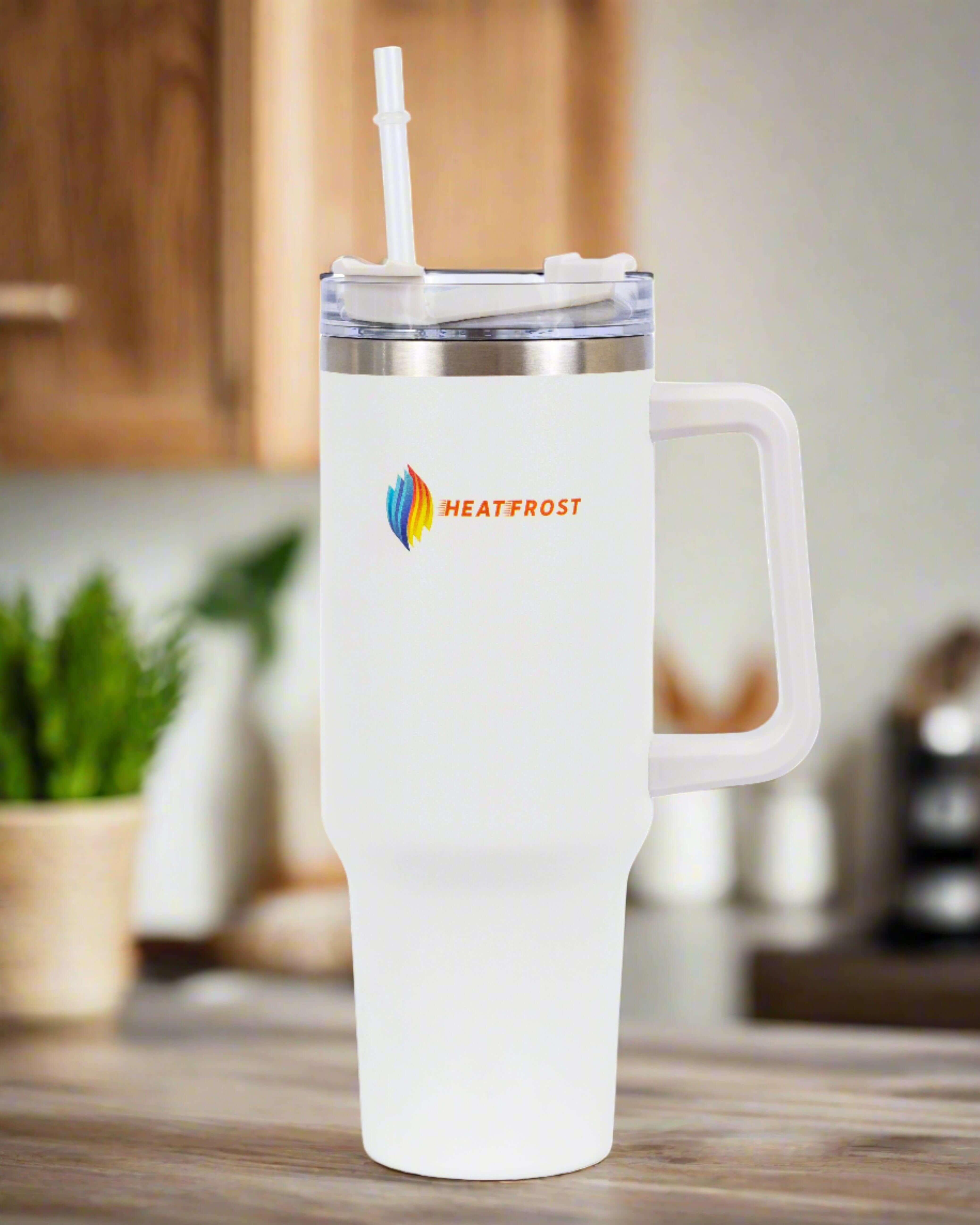 A 40oz stainless steel tumbler, designed as a travel-friendly quencher and adorned with the "Heatfrost" logo in orange, rests on a kitchen counter. This insulated beverage container features a white exterior and comes with a transparent lid and straw. In the softly blurred background, wooden cabinets and a green plant can be seen. It's an essential accessory for drink enthusiasts!