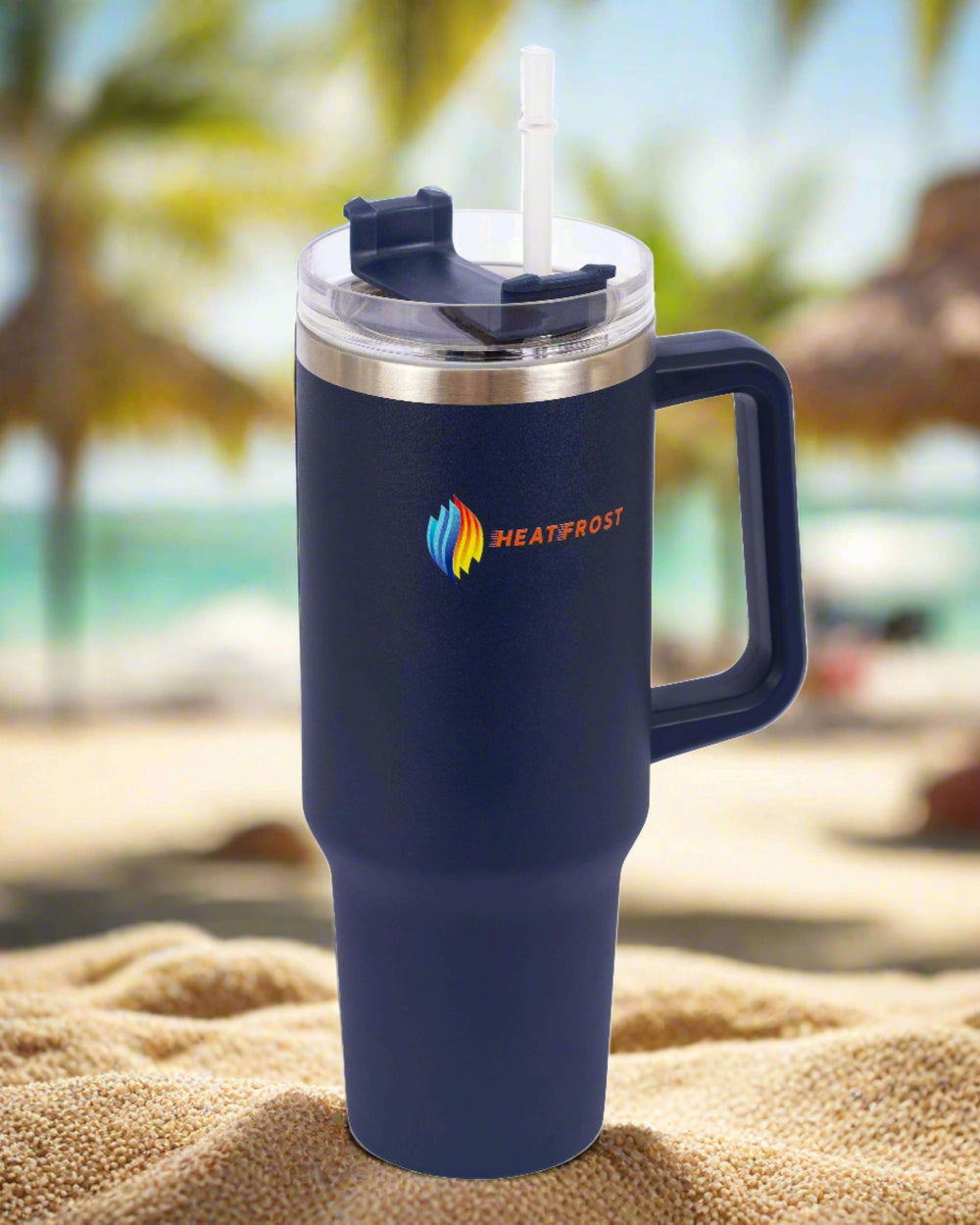 A "HEATFROST" logo-adorned 40oz Stainless Steel Tumbler Insulated Beverage Container, featuring a handle and straw, rests on the sandy ground. This travel-friendly quencher is perfect for those who love to keep their drinks at the ideal temperature. A blurred beach with palm trees in the background hints at a tropical locale.