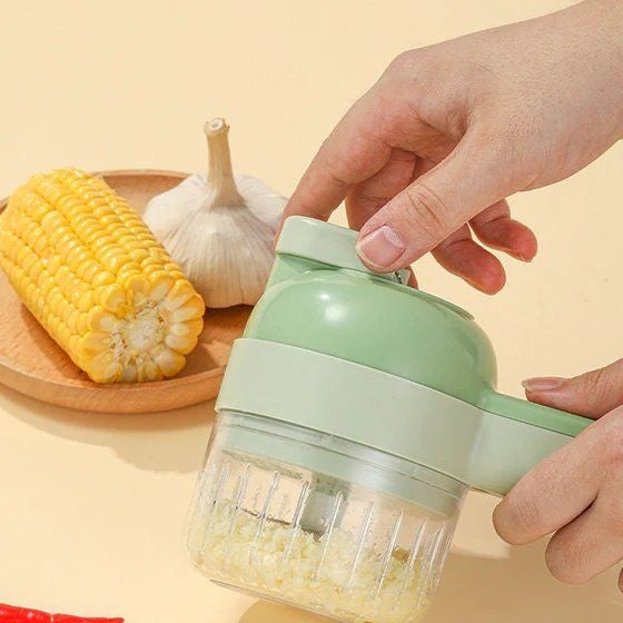 A person skillfully operates a green multifunctional electric vegetable slicer as they mince garlic, prominently featured in the scene. Meanwhile, a wooden plate adorned with half a cob of corn and a garlic bulb serves as decoration in the background. This unfolds on a light-colored surface, highlighting the innovative design of the Vegetable Slicer that seamlessly blends traditional aesthetics with modern functionality.