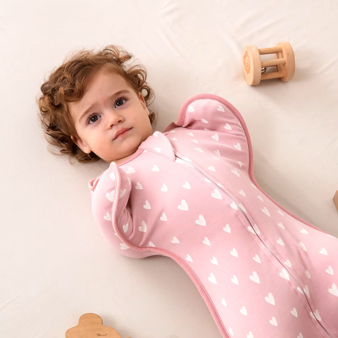 A baby with curly hair lies on a soft surface, swaddled in a Baby Sleeping Bag Newborn Baby Swaddle Summer Cotton Sleep Sack with pink fabric adorned with white heart patterns. Wooden toys are scattered nearby, and the baby gazes up calmly, enjoying the peaceful sleeping environment.