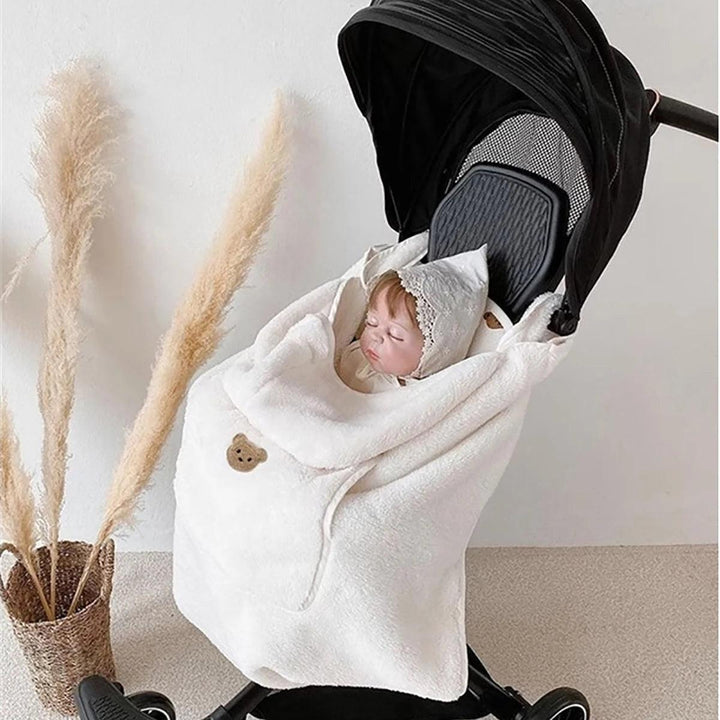 A baby sleeps peacefully in a black stroller, wrapped warmly in the cozy Baby Stroller Windproof Cover—a white, bear-themed blanket perfect for autumn and winter. The serene and minimalistic setting is complemented by a decorative vase with pampas grass.