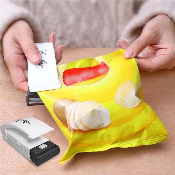 A person is using a small, white Heat Bag Sealer to close a yellow chip bag with a transparent window, placed on a wooden table. The bag features images of sliced chips. Another Food Packaging Sealing Machine lies nearby, ensuring all snack bags stay fresh.