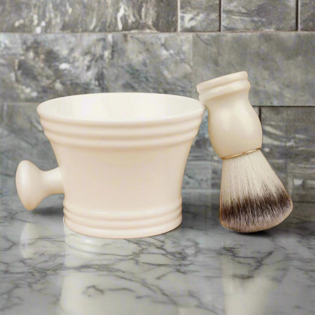 A cream-colored shaving bowl with a side handle rests on a marble surface. Next to it, the Beard Shaving Brush Wooden Handle Bristle Hair Brush, featuring white and brown bristles and a sleek wooden handle, adds elegance to the scene. The gray tiled marble wall in the background enhances this timeless grooming setup.