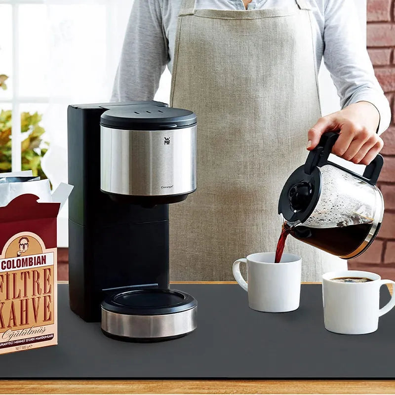 A person in an apron is pouring coffee from a glass carafe into two white mugs beside a black and silver coffee maker. On the counter, next to the box labeled "Colombian Filtre Kahve," lies a "Super absorbent kitchen mat" with an anti-slip texture, ensuring quick drying in this cozy kitchen setting.