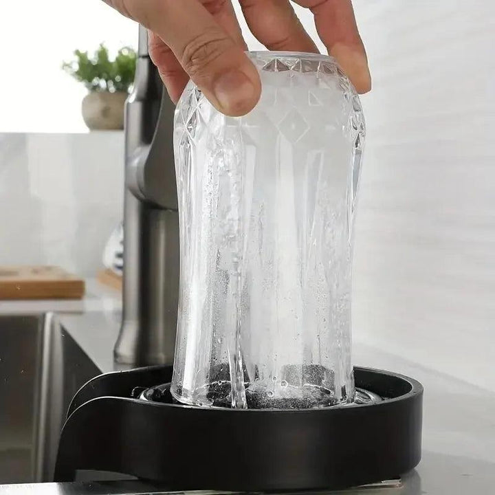 A hand holds an upside-down glass being rinsed on the Automatic Cup Washer High Pressure Glass Cleaner, which is installed on a black countertop. Water sprays upward, efficiently cleaning the glass. A faucet and modern kitchen decor are visible in the background.