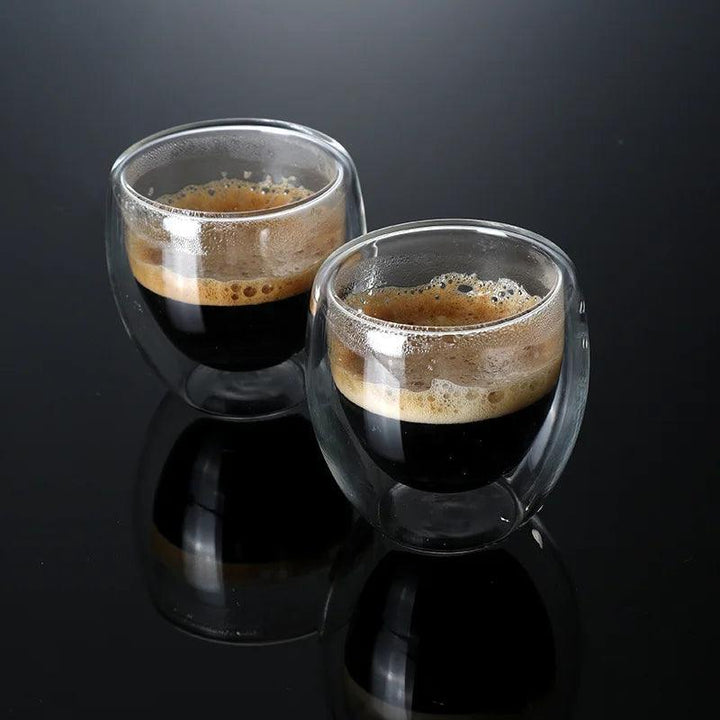 Two Double-layer insulated glass cups, each with an 80ml capacity, filled with espresso sit on a glossy black surface. The coffee has a rich, dark color topped with a layer of light brown crema, and the reflection of these elegant cups is visible beneath their form.