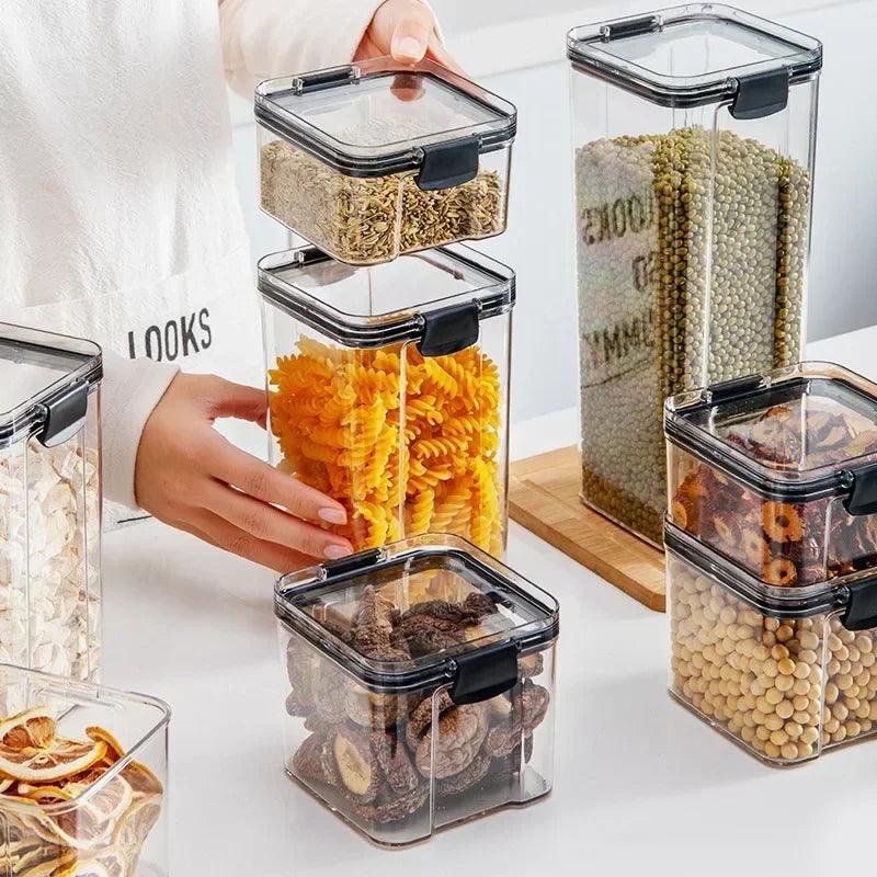 A person arranges Plastic Food Containers filled with various dry foods, such as pasta, seeds, mushrooms, and nuts, on a white kitchen counter. These airtight containers, topped with black lids and stacked neatly, are perfect for storing food items efficiently.