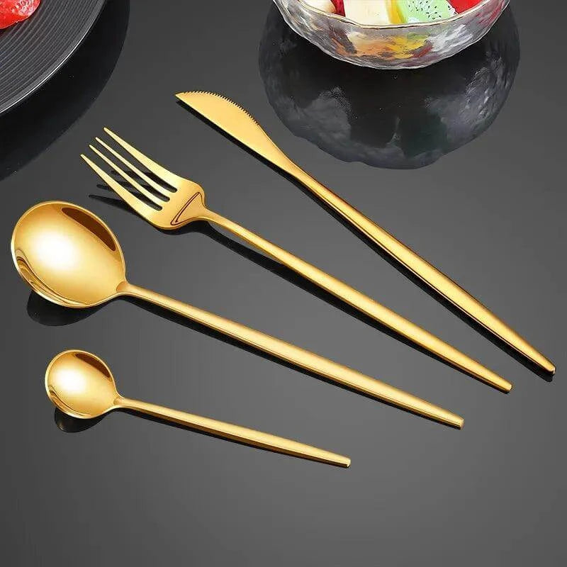 A set from the Gold Dinnerware Set Stainless Steel Flatware Elegant Table Setting, featuring a stunning knife, fork, tablespoon, and teaspoon in gold-colored stainless steel, is elegantly arranged on a glossy black surface. In the background, a glass bowl filled with colorful contents adds a playful touch to this sophisticated display.