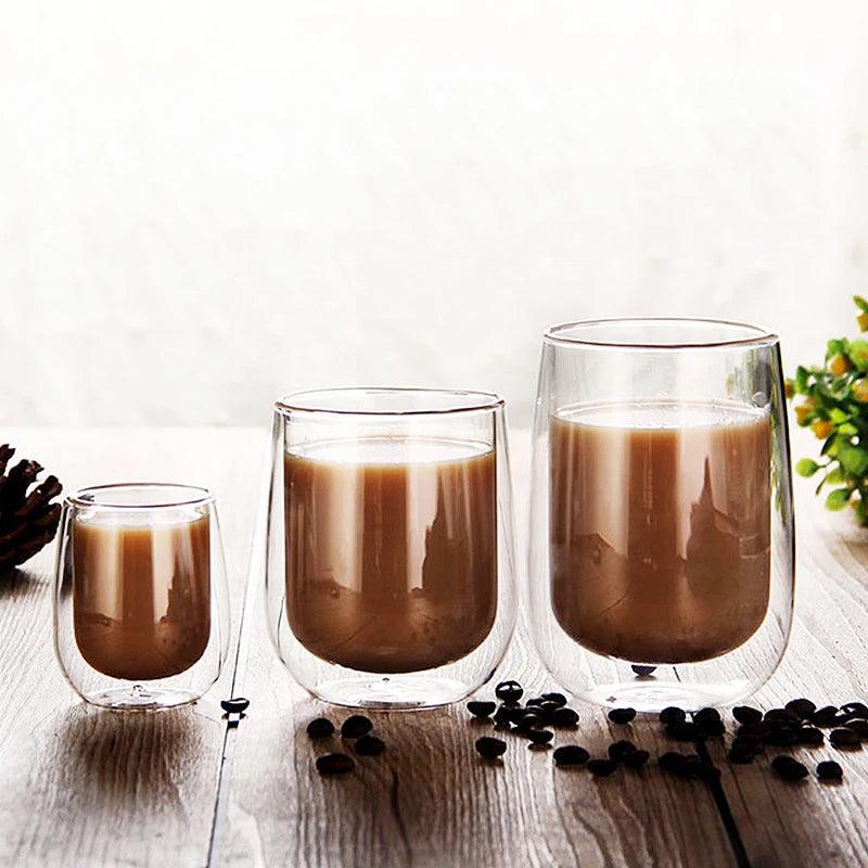 Three Clear Double Wall Glass Coffee Mugs, each of a different size and filled with a brown beverage, sit on a wooden table. Nearby, coffee beans are scattered to enhance the eco-friendly vibe, while a green plant and pine cone contribute to the natural backdrop.