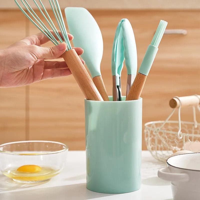 A hand reaches for a whisk in a mint green utensil holder filled with items from the Silicone Cooking Utensils Non-Stick Kitchen Tools BPA-Free Cooking Set, featuring eco-friendly tools like a spatula, tongs, and a basting brush. In the background on a wooden countertop are a bowl with a cracked egg and a wire basket.