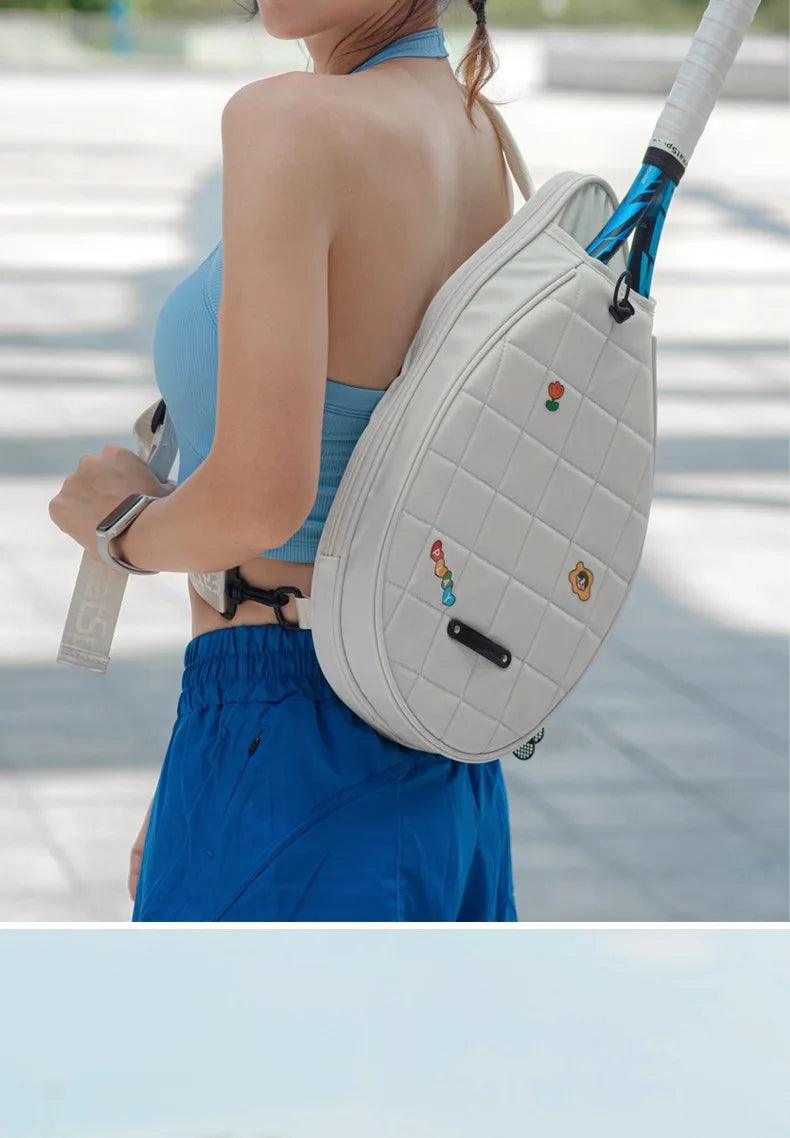 A woman wearing a blue top and shorts carries the Four-Slam Tennis Bag, Badminton Pickleball Bag, Korean Style Sports Bag, which is quilted and white with small, colorful pins, slung over her shoulder. She holds two rackets while standing outdoors on a bright, sunny day.