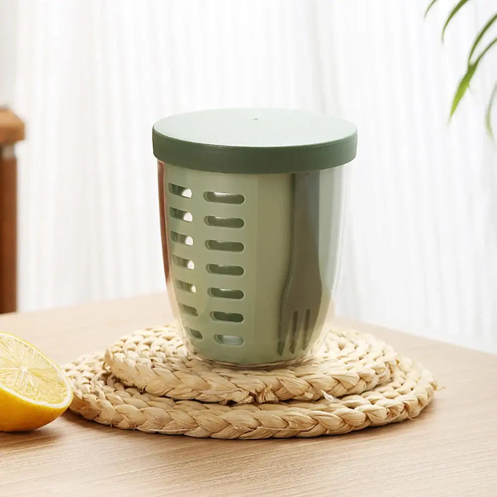 A green "Fruit Cup with Fork Portable Picnic Bottle" rests on a woven mat on a wooden table. To the left, a sliced lemon adds a splash of color, while sheer curtains in the background allow natural light to emphasize this eco-friendly kitchen essential.