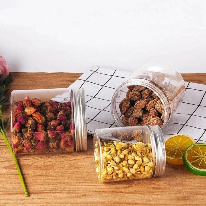 Three Clear Storage Jars Empty Cosmetic Containers Travel Bottles are placed on a wooden table, each filled with an assortment of dried items: red rose buds, light yellow flowers, and brown nuts or roots. Nearby, a checkered cloth complements sliced lemon and lime, while a pink flower adds decoration to the corner.