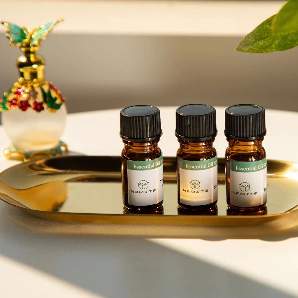 Three small bottles of "Natural plant essential oil" are placed on a golden tray. A decorative perfume bottle with a colorful design stands in the background. Soft lighting and a touch of green foliage create a serene atmosphere, perfect for aromatherapy.