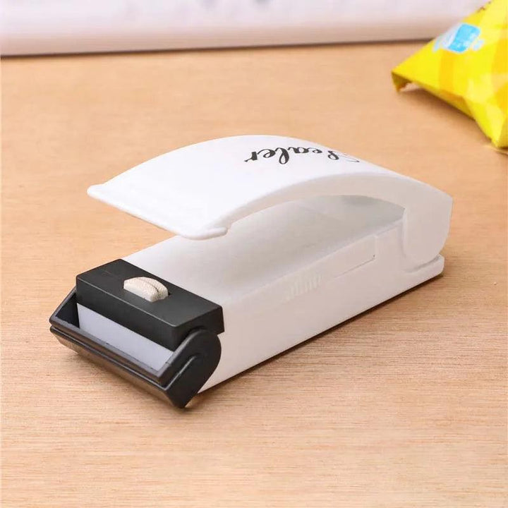 A white "Heat Bag Sealer, Food Packaging Sealing Machine" designed for sealing food storage bags rests on a wooden table beside the corner of a yellow snack bag. This portable device showcases a curved handle and a black sealing element at the front.