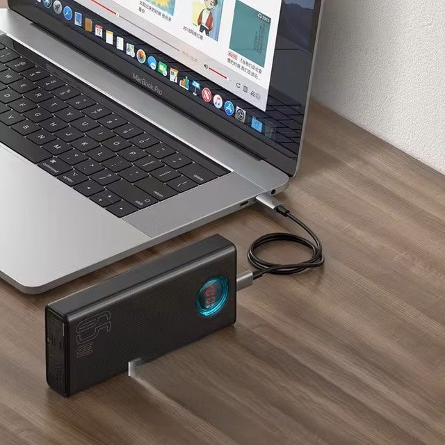 A laptop rests on a wooden desk, connected to a 30000mAh portable external power bank with 65W PD3.0 quick charging capability via a cable. The power bank's digital display indicates the charge percentage, while the laptop screen is vibrant with various apps and windows open.