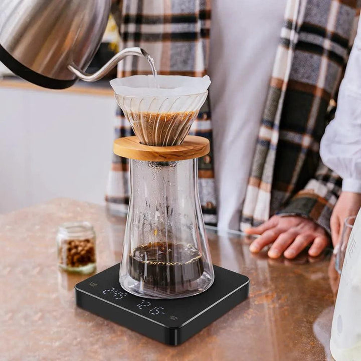 A person carefully pours hot water from a kettle into a glass pour-over coffee maker, sitting atop the Digital Coffee Scale with Timer High Precision Coffee Scale USB Rechargeable Coffee Scale. The setup adds elegance to the kitchen counter, with coffee beans in a jar enhancing the scene's aroma and allure.