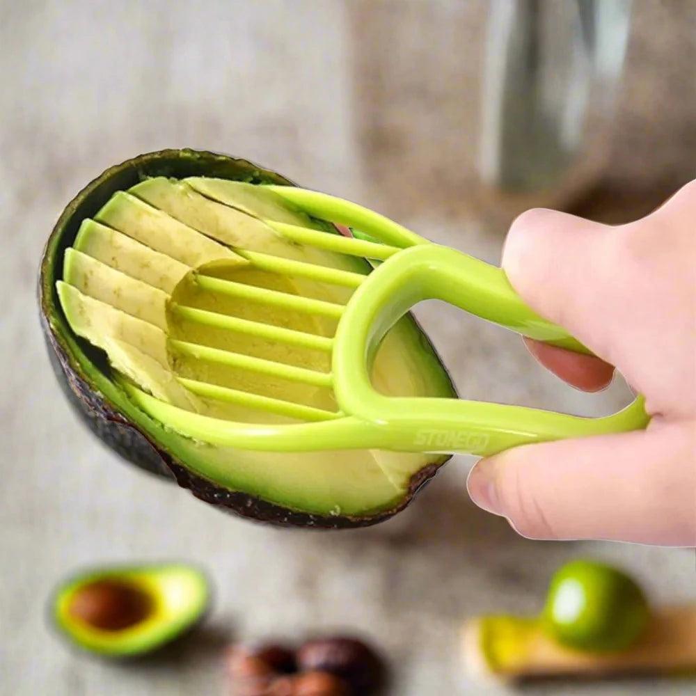 Using the Non-Toxic 3-in-1 Avocado Slicer Knife, a hand expertly slices even pieces from a halved avocado. This efficient fruit peeler and pulp separator, an essential kitchen tool, is highlighted against a backdrop of blurred avocado halves and other kitchen items.