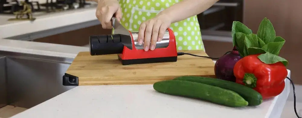 Person using an electric knife sharpener on a wooden cutting board with fresh vegetables.