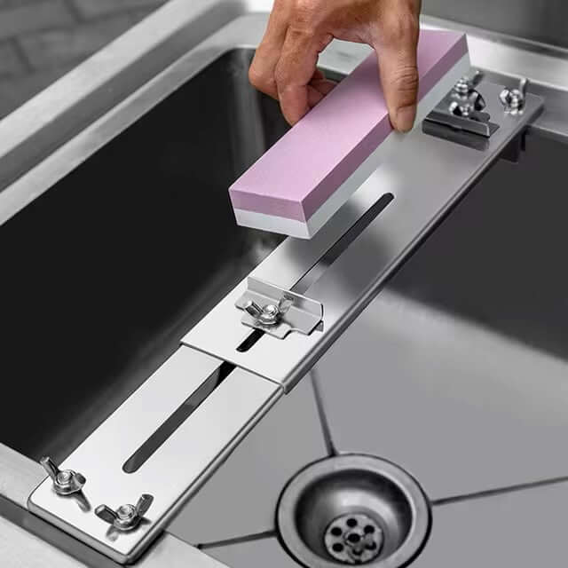 A hand adjusts the Whetstone Knife Sharpener, featuring an attached pink sharpening stone, on a stainless steel sink. This eco-friendly sharpener includes a Water Stone Plate and Adjustable Whetstones Sink Bridge Holder, enhancing the sleek design of the sink with its visible drain hole.