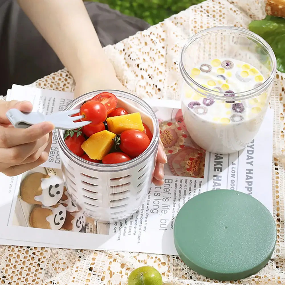 A person holding a fork over a Fruit Cup with Fork Portable Picnic Bottle, filled with cherry tomatoes and cheese cubes. Next to it is another Fruit Cup used as a strainer for cereal and milk, ensuring spill-free storage. A green lid and a printed fabric are also visible on the surface.