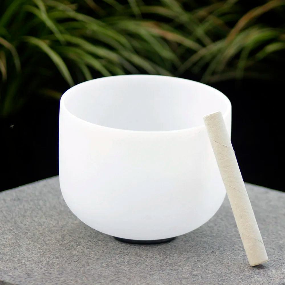 A Note B Chakra Meditation and Yoga Bowl rests alongside a mallet on a gray stone surface, surrounded by softly blurred green plants. Perfect for sound healing, its soothing tones offer tranquility and harmony.