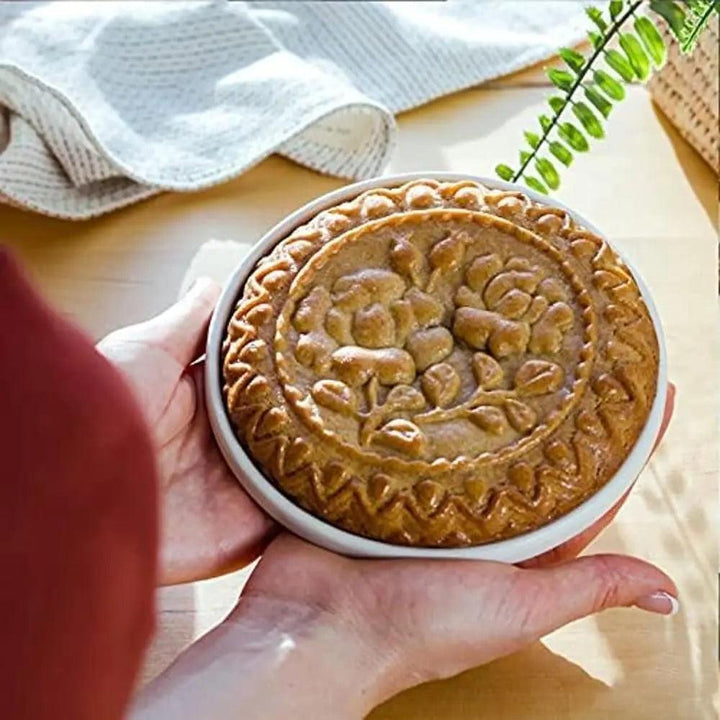 A person holds a round pie with intricate floral patterns on its crust, reminiscent of designs made with the Wooden Shortbread Cookie Mold Valentine's Day. The pie is placed on a white plate, and sunlight highlights its golden-brown surface. A fern can be seen in the background.