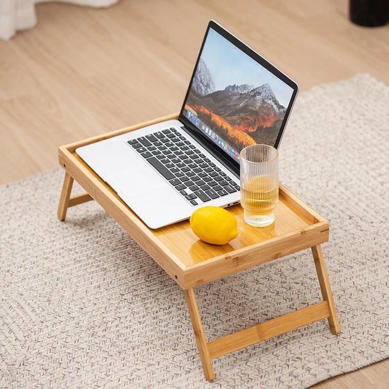 A Bed Tray Table with Folding Legs, crafted from bamboo and ideal as a home workspace accessory, is positioned on a textured rug. It supports an open laptop alongside a clear glass of water and a yellow lemon, all atop a light-colored wooden floor in this welcoming setup.