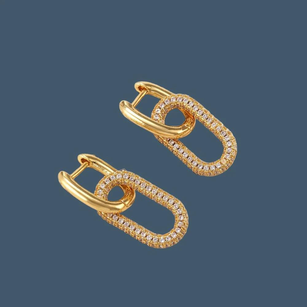 A pair of elegant gold-plated "Trendy U-shape earrings" are displayed on a blue background, each adorned with a geometric design encrusted with sparkling CZ gemstones. These "Fashion CZ earrings" are perfect for adding a touch of sophistication to any outfit.