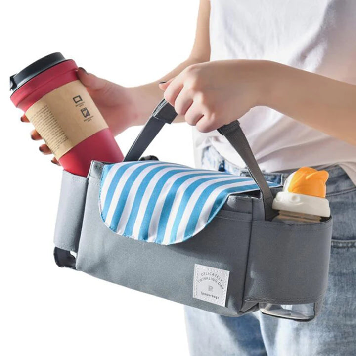 Someone is using a "Stroller bag organizer," which is crafted from durable nylon fabric in gray and features a blue and white striped flap. Inside, there's room for essentials like a red travel coffee mug and a baby bottle, providing ample storage space. Additionally, the organizer includes a handle for easy carrying.