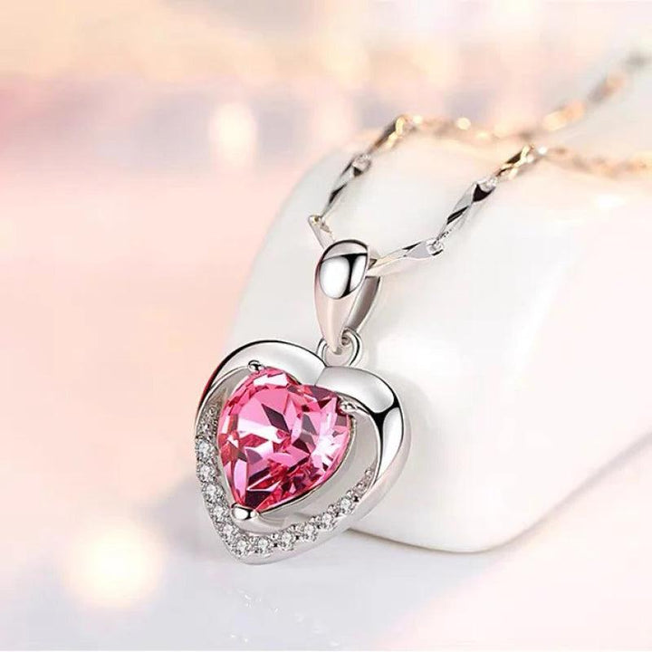 The 925 Silver Pendant Necklace Pink Heart Crystal Hypoallergenic Jewelry showcases a shimmering pink heart crystal, beautifully accented with petite cubic zirconia diamonds along its edges. This piece gracefully dangles from a delicate chain, set against a softly illuminated and gently blurred backdrop.