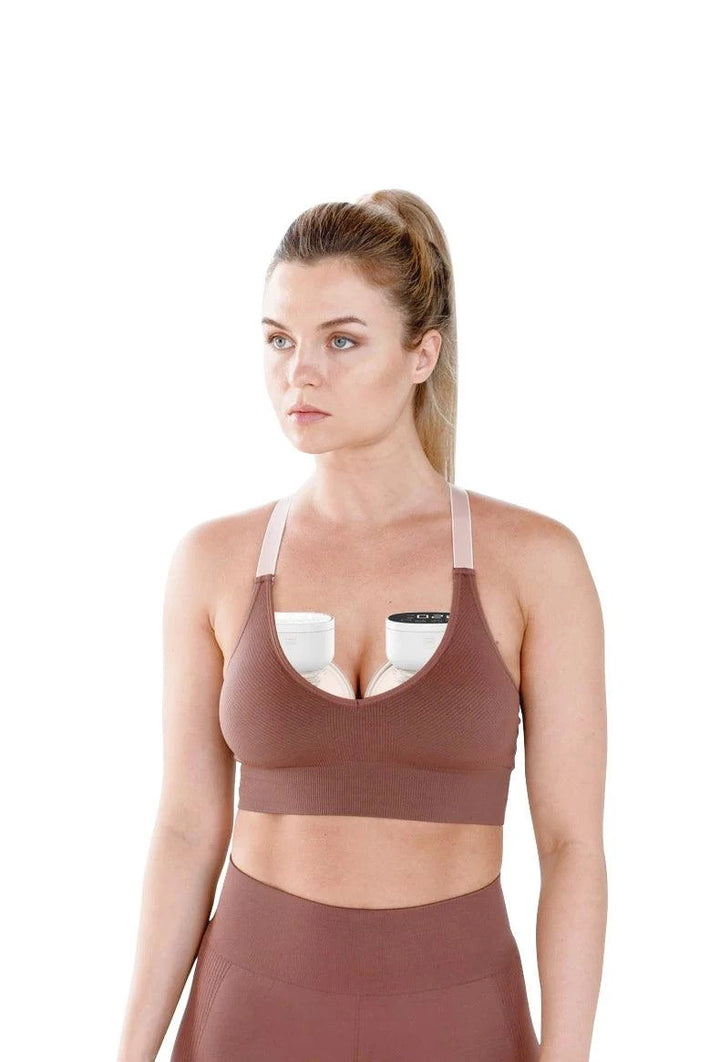 A new mom with her hair in a ponytail, sporting a brown sports bra, looks off-camera. Discreetly tucked inside is an Automatic, Hands-Free Breast Pump, blending comfort and practicality against the plain white background.