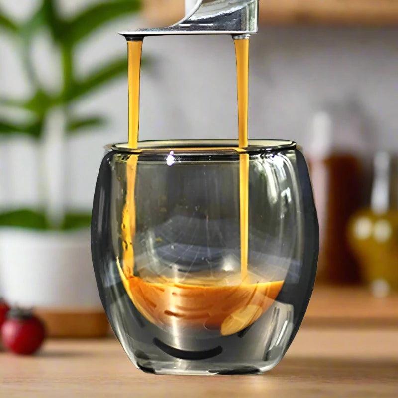 Espresso gracefully flows from the coffee machine into a double-layer insulated espresso glass, part of a set ranging from 2 to 6 pieces with an 80ml capacity, resting on a wooden surface. A blurred green plant and various kitchen items enhance the charm of the background while preserving the optimal temperature for your perfect brew.