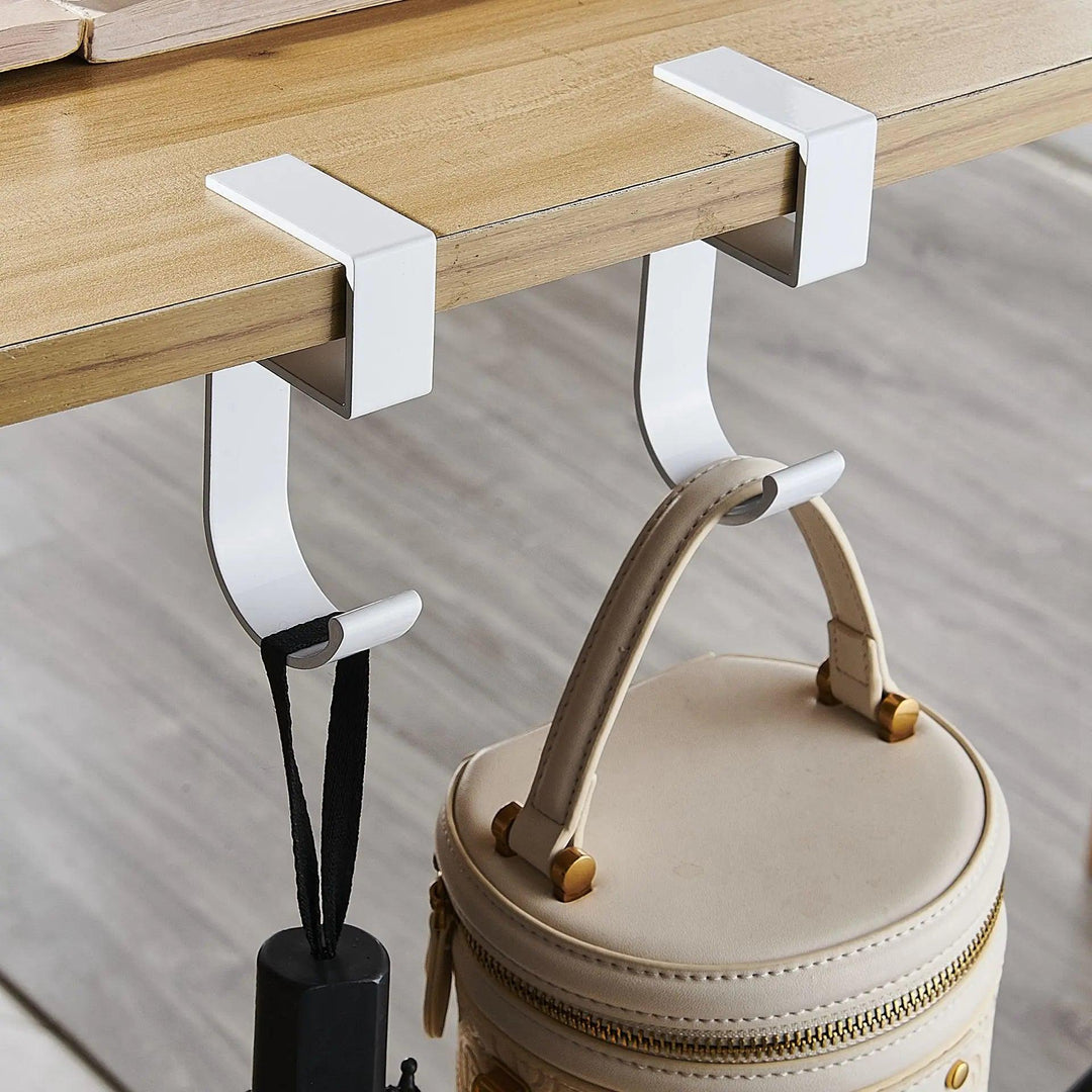 White metal hooks with a minimalist design are attached to a wooden table edge, securely holding a tan leather bag and a black item. This sleek "Portable hanging bag hook" or "Desk side table hook" is perfect for keeping your essentials within reach as a removable handbag holder.