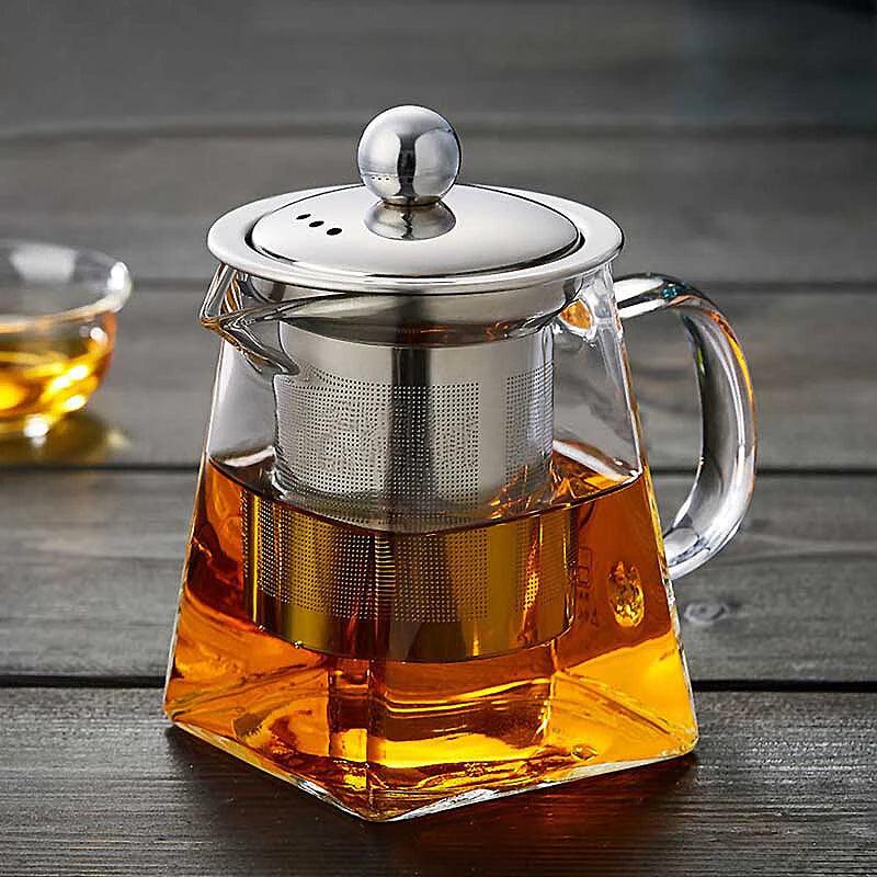 The Heat Resistant Glass Teapot with a Stainless Steel Tea Infuser Filter Flower, filled with amber-colored tea, sits on a dark wooden surface. In the background, a small shallow glass bowl holds some tea.